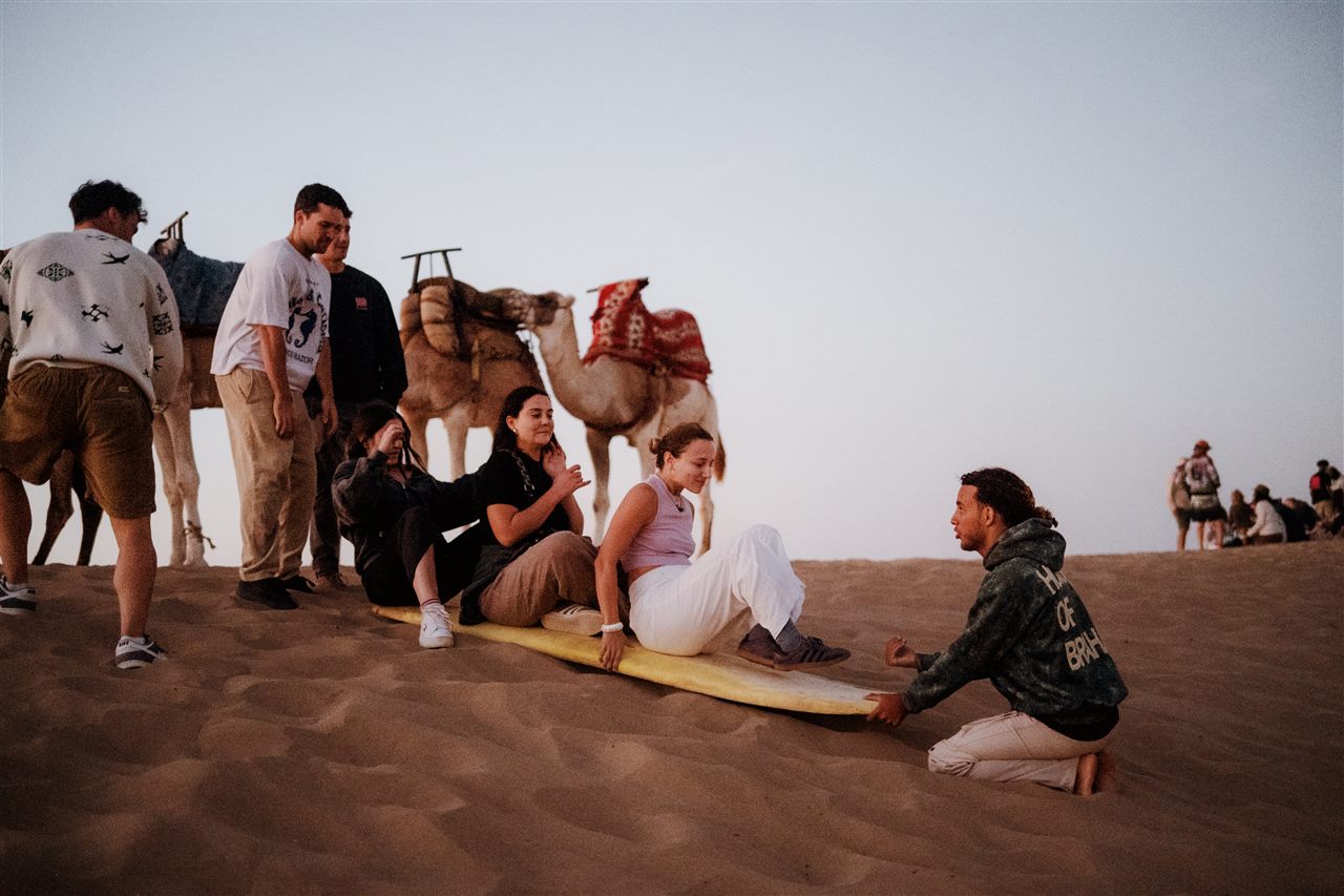Group sandboarding with traditional Moroccan camels on the dunes near Taghazout